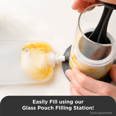 Hands using a glass pouch filling station to fill a clear glass pouch with a black plunger, with a translucent squeeze pouch on a white surface and a text banner at the bottom.