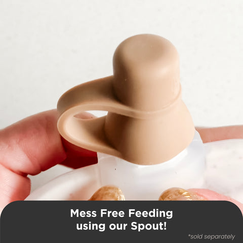 A close-up of a hand holding a beige silicone baby feeding spout over a white feeding tray, with a dark text banner at the bottom reading 'Mess Free Feeding using our Spout!'.