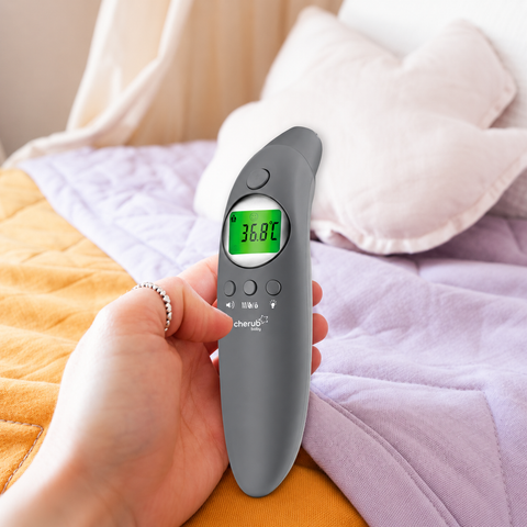 Hand holding a digital thermometer with a blurred background of a bed.