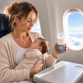 Mother holding her baby on an airplane while holding a portable bottle warmer V3 with a baby bottle attached.