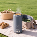 A matte gray smart water bottle with a digital temperature display and steam vent, shown outdoors on a picnic blanket next to a woven basket, pastries, and a beige lunch bag.